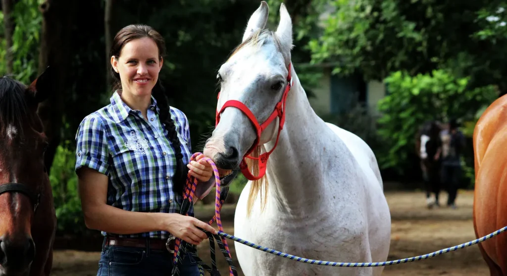 A woman in a plaid shirt stands with a grey horse wearing a red halter. Two other horses are visible in the background.