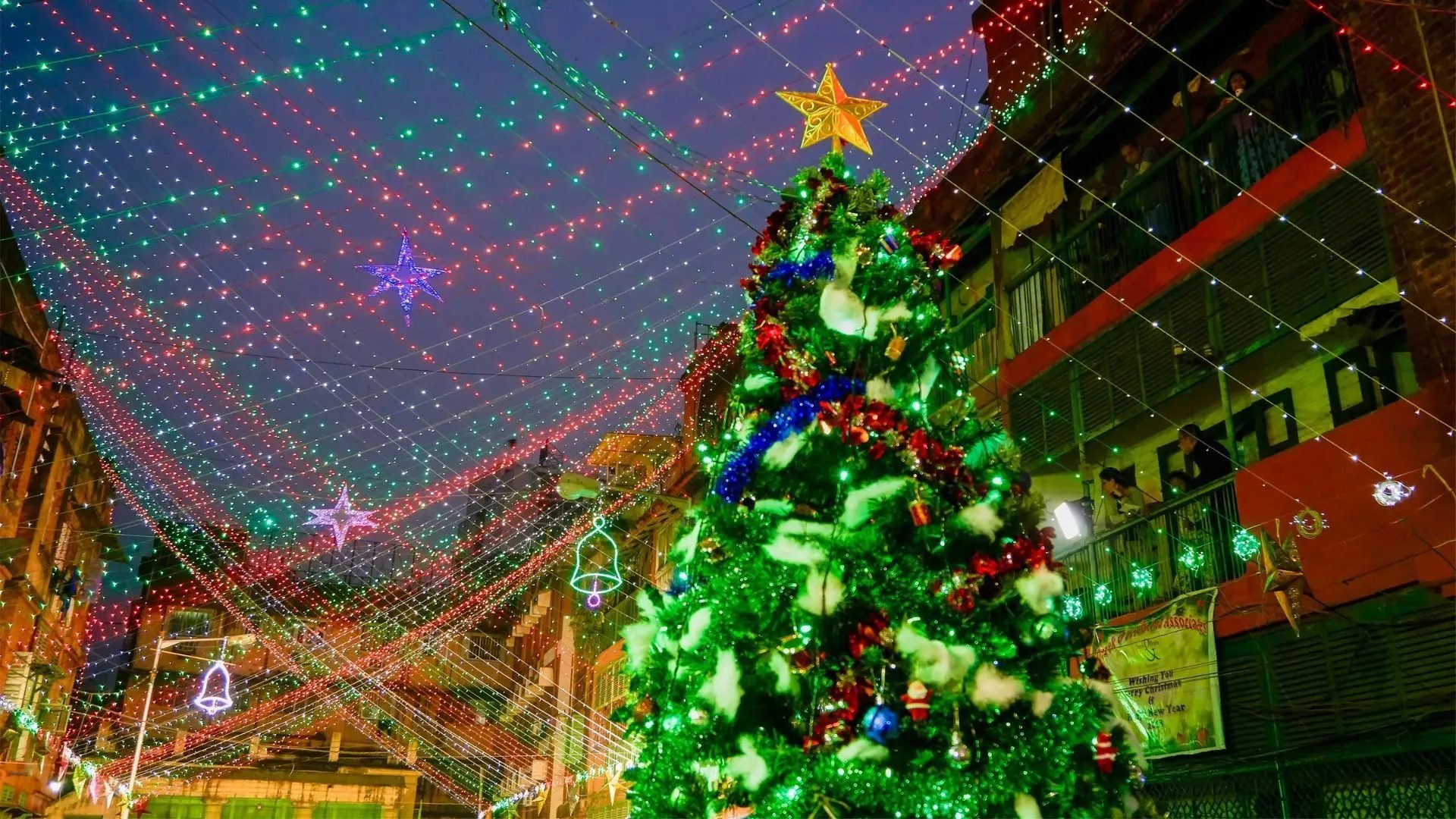 Christmas tree decorated with colorful ornaments and lights, standing under festive string lights in a bustling street at dusk.