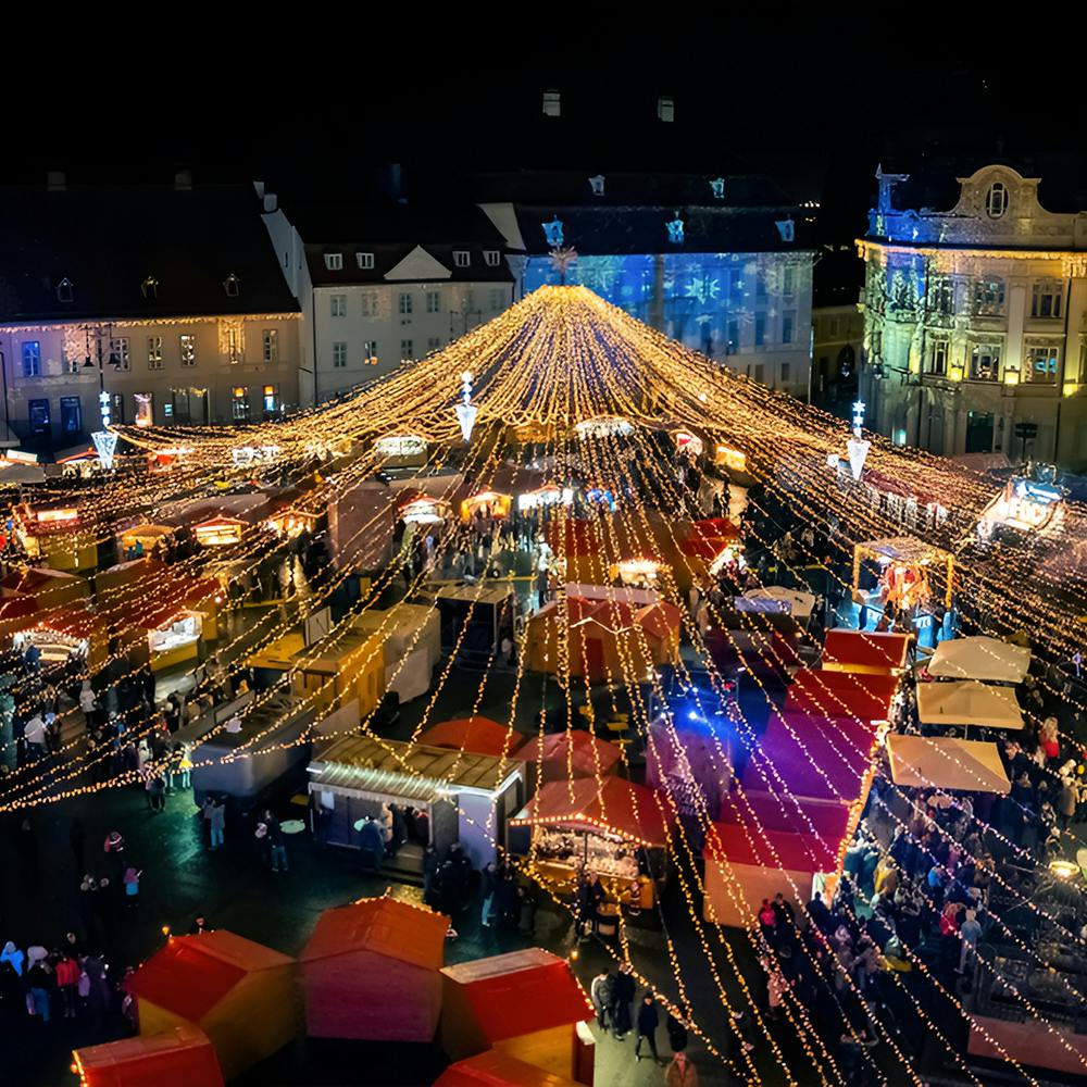 Aerial view of a festive nighttime market square decorated with illuminated string lights, surrounded by buildings and bustling with people visiting various stalls.