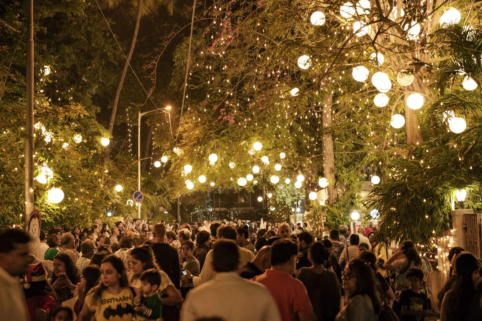 Crowded night market or street festival illuminated with glowing string lights hanging from trees, surrounded by people enjoying the vibrant outdoor event