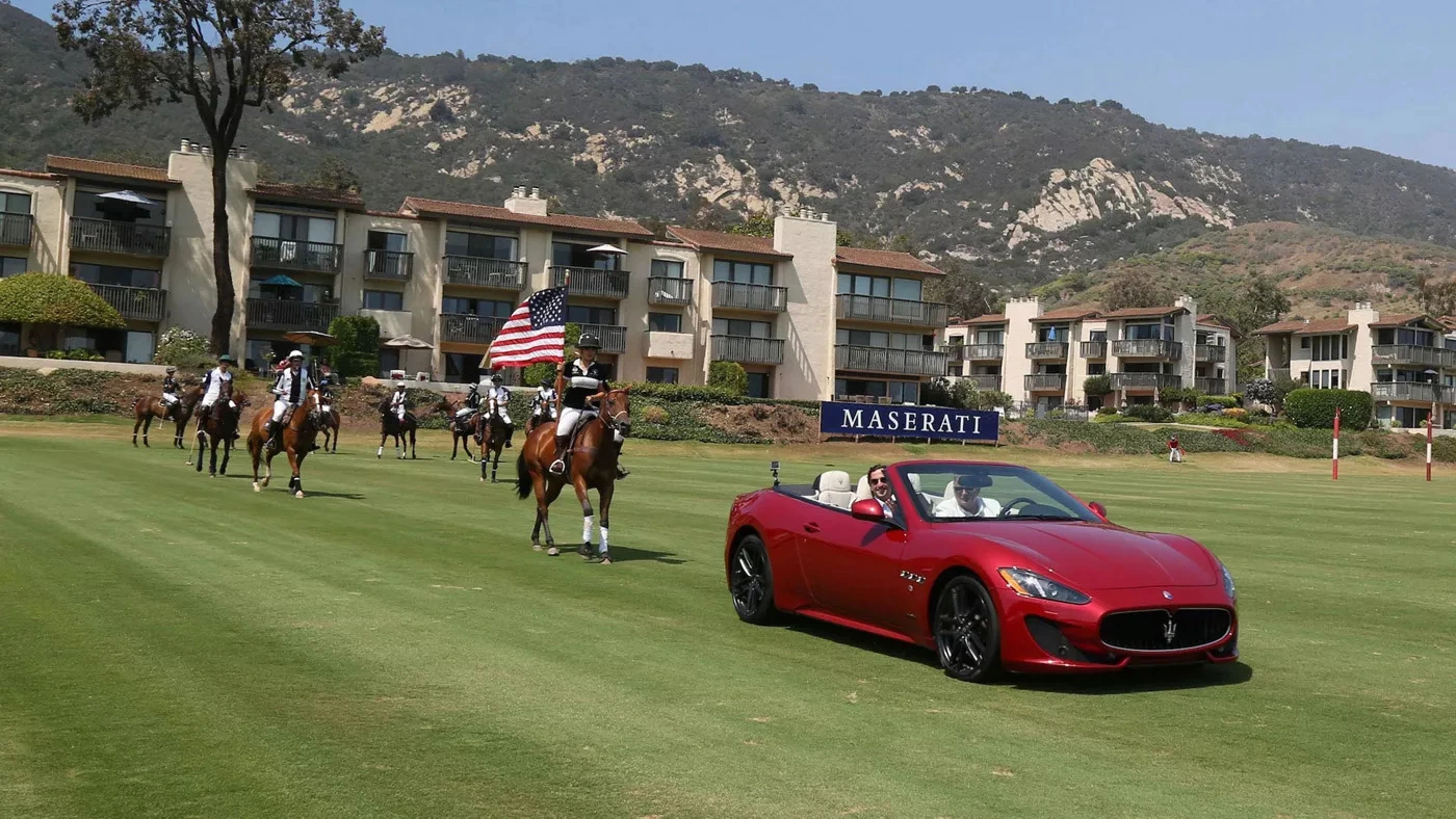 Parade of horseback riders carrying American flags, followed by a red Maserati on a polo field – a vibrant display of luxury and sport