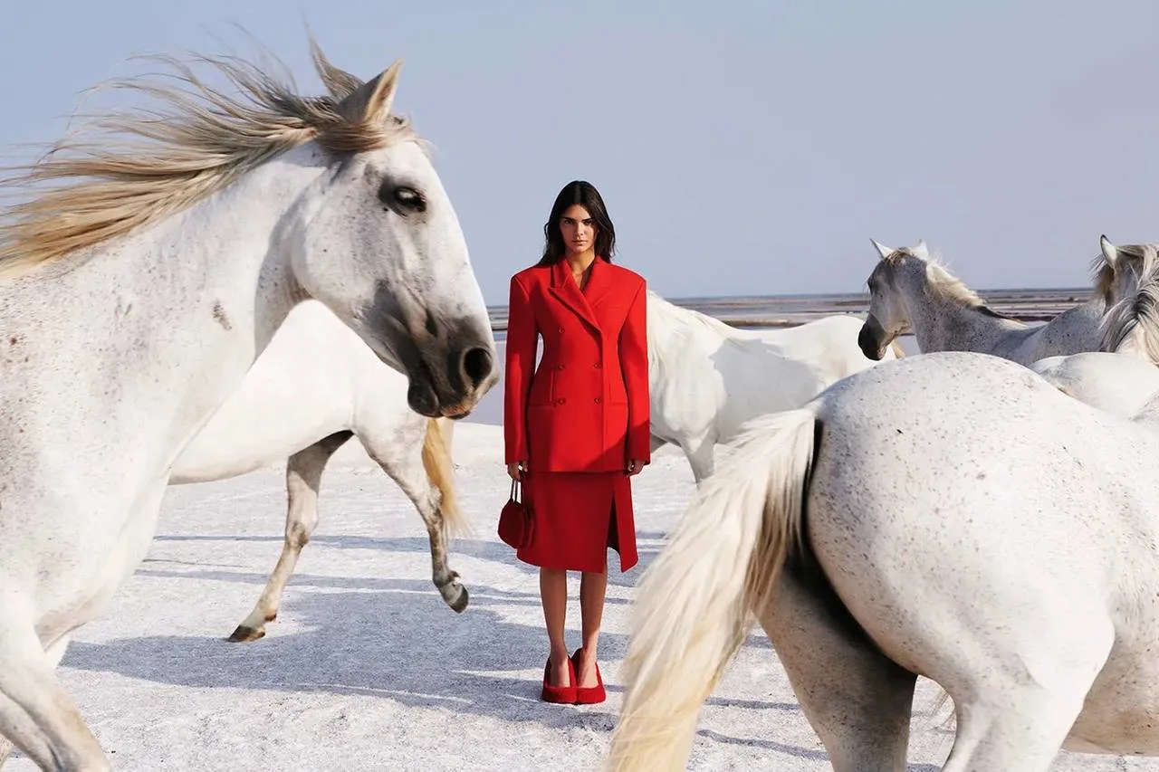 A woman wearing a vibrant red dress poses elegantly before several horses in a picturesque landscape
